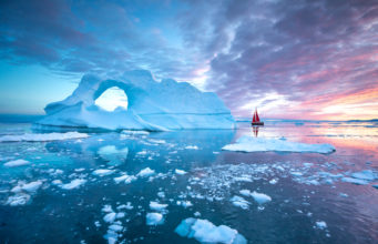 Little red sailboat cruising among floating icebergs in Disko Bay glacier during midnight sun season of polar summer. Ilulissat, Greenland.