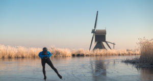 Person ice skating alone on a frozen river past a windmill and hoar frosted reeds in Kinderdijk in winter in the Netherlands.